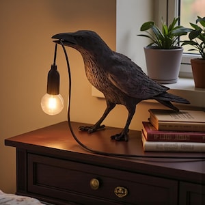 Könnte beinhalten: Dunkelgraue, rabenförmige Lampe mit einer Glühbirne im Schnabel. Die Lampe steht auf einem dunkelbraunen Nachttisch mit einer Schublade. Das Kabel der Lampe ist schwarz. Die Lampe steht neben einem Stapel Bücher und einem Fenster mit Pflanzen.