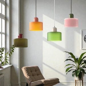 May include: Four contemporary pendant lights in orange, green, pink, and olive green. Each light has a cylindrical shade and a contrasting top. The lights hang from the ceiling in a bright room with a chair and plants.