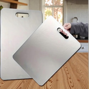 以下が含まれることがあります： 丸みを帯びた角を持つ2つのステンレス鋼製の長方形まな板。1つは持ち上げられており、ハンドルの切り欠きが見えます。まな板は木製の表面に置かれており、キッチンを連想させます。