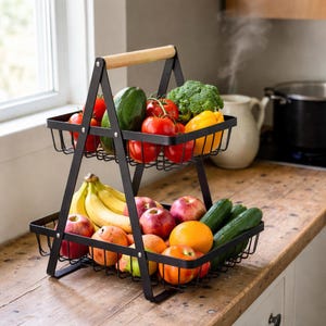 May include: A two-tiered black metal wire basket filled with fresh produce. The top tier holds tomatoes, avocado, broccoli, and yellow bell peppers. The bottom tier contains bananas, apples, oranges, and cucumbers. A wooden handle is at the top.