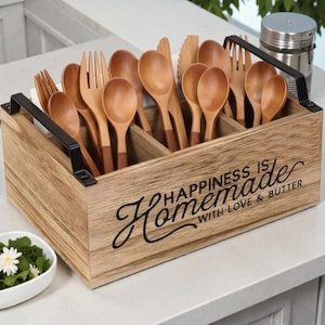 K&ouml;nnte beinhalten: Holzbesteckhalter mit schwarzen Griffen, gef&uuml;llt mit Holzl&ouml;ffeln, Gabeln und Messern. Der Halter tr&auml;gt den Text "Happiness is Homemade with Love & Butter". Das Besteck ist hellbraun.