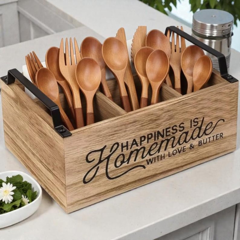 K&ouml;nnte beinhalten: Holzbesteckhalter mit schwarzen Griffen, gef&uuml;llt mit Holzl&ouml;ffeln, Gabeln und Messern. Der Halter tr&auml;gt den Text "Happiness is Homemade with Love & Butter". Das Besteck ist hellbraun.