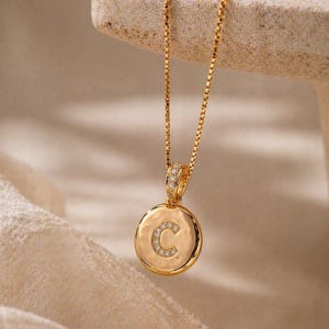 Könnte beinhalten: Goldfarbene Halskette mit einem Anhänger mit dem Buchstaben "C", der mit kleinen, klaren Edelsteinen besetzt ist. Der Anhänger ist rund und hat eine gehämmerten Textur. Die Kette ist filigran und ebenfalls goldfarben.