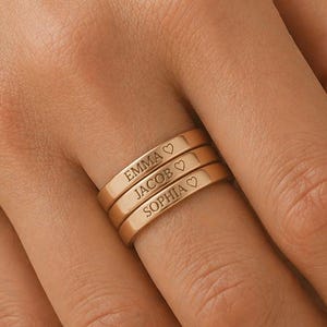 Könnte beinhalten: Drei goldfarbene Ringe, die auf einem Finger gestapelt sind. Jeder Ring ist mit einem Namen und einem Herzsymbol graviert: Emma, Jacob und Sophia. Die Ringe werden auf einer Hand mit neutralem Hintergrund gezeigt.