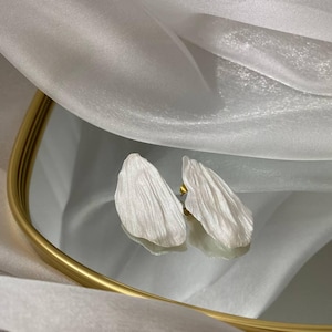 Könnte beinhalten: Ein Paar weiße, blütenblattförmige Ohrringe mit goldfarbenen Stiften. Die Ohrringe werden auf einer reflektierenden Oberfläche in einem goldgerahmten Tablett mit weißem Stoffhintergrund präsentiert.