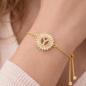 Könnte beinhalten: Goldfarbenes Armband mit einem kreisförmigen Anhänger, der einen Buchstaben "Y" in der Mitte und klare, baguetteförmige Kristalle aufweist. Das Armband hat eine feine Kette und einen Schiebeverschluss mit Kugeln zur Größenanpassung.