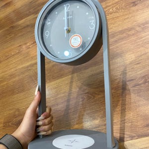 Peut inclure: Une horloge de bureau grise avec un cadran circulaire et un cadre incurvé en forme d'arche. Le cadran affiche l'heure avec des chiffres et des aiguilles noirs. La base est ronde avec un bouton et le mot "Athens" est visible.