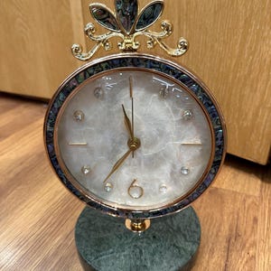 Op de afbeelding: Een decoratieve tafelklok met een ronde wijzerplaat, een witte marmerachtige achtergrond en gouden wijzers. De klok is versierd met kristallen accenten en een goudkleurig frame. De basis is groen marmer en de bovenkant heeft een decoratief goud- en abalone schelpontwerp.