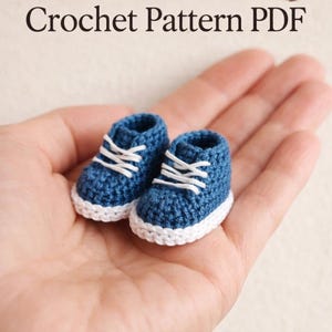 May include: A pair of tiny, handmade blue crochet sneakers with white laces and soles. The text "Crochet Pattern PDF" is at the top of the image. These miniature shoes are perfect for crafting projects or as a unique decorative item.