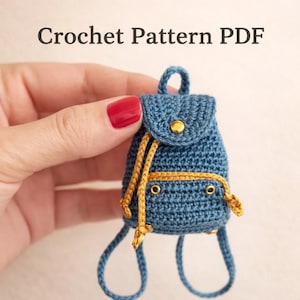 May include: A miniature blue crochet backpack with gold accents, including a drawstring and a button closure. The text "Crochet Pattern PDF" is at the top. The backpack has two blue loops at the bottom.
