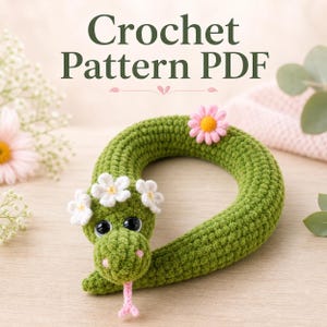 Peut inclure: Un serpent vert crocheté orné de fleurs blanches et roses. Le serpent a de grands yeux noirs et une langue rose. Le texte "Crochet Pattern PDF" est visible en haut de l'image.