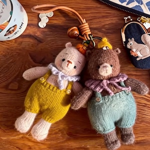 May include: Two small, knitted bear keychains attached to a leather strap. The left bear is cream-colored with yellow overalls and a light purple ruffled collar. The right bear is brown with teal overalls and a lavender ruffled collar.