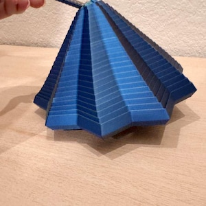May include: A blue, geometric paper sculpture with a layered, fan-like design. The sculpture is held by a thin, dark handle. The color gradient ranges from light to dark blue, creating a three-dimensional effect. The object is displayed on a light wooden surface.