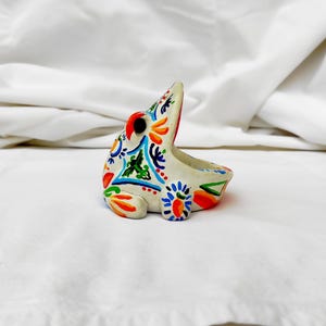 Puede incluir: Pequeña figura de rana de cerámica blanca con vibrantes diseños florales pintados a mano en rojo, naranja, azul y verde. La rana tiene la boca abierta y probablemente sea un objeto decorativo o un pequeño macetero.