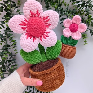 以下が含まれることがあります： 茶色の鉢植えに2つの花が咲くかぎ針編みのフラワーアレンジメント。大きな花は、赤いアクセントと赤い中心部を持つピンク色の花びらを持っています。小さな花はピンク色で、緑色の茎と葉があります。鉢は茶色です。