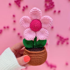 以下が含まれることがあります： 手作りのかぎ針編みの花が茶色の鉢に入っています。花はピンク色の花びらと濃いピンク色の中心部を持っています。緑の葉が花の下に見えます。鉢は茶色です。背景はピンク色です。