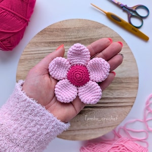 以下が含まれることがあります： ピンクの花びらと濃いピンクの中心部を持つ手作りのかぎ針編みの花。 花は手で持たれており、近くに糸、はさみ、かぎ針編みが置かれています。 花は木製の表面にあります。 テキスト「Tambo_crochet」が表示されます。