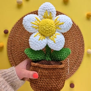 以下が含まれることがあります： 手作りのかぎ針編みの花、茶色の鉢植え。 花は白い花びらと黄色のアクセント、黄色の中心部があります。 緑の葉と茶色の茎がデザインを完成させています。 鉢は茶色で、質感があります。 背景は黄色です。