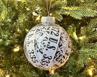 May include: A clear glass Christmas ornament with a silver top and a white ribbon wrapped around it. The ribbon has black numbers printed on it, including 2, 3, 5, 7, 11, 13, 17, 19, 23, 29, 31, 37, 41, 43, 47, 53, 59, 61, 67, 71, 73, 79, 83, 89, 97.