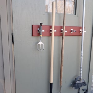 Puede incluir: Una colección de herramientas de jardinería colgadas en un estante de madera rojo fijado a una puerta de cobertizo verde. Las herramientas incluyen una horquilla de jardín, una azada y una escoba. Las herramientas tienen componentes de madera y metal.