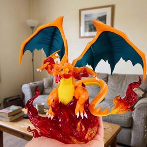 May include: A detailed Charizard figurine, a popular character from the Pokémon franchise. The figure is orange and yellow with blue wings, posed on a base of red flames. A collectible item, it is likely made of resin or plastic.