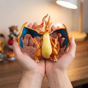 Könnte beinhalten: Eine Charizard-Figur, eine beliebte Figur aus dem Pokémon-Franchise, wird in geformten Händen gehalten. Die Figur hat orange, gelbe und blaue Farben. Die Flügel sind leuchtend blau mit orangefarbenen Rändern. Die Figur ist detailliert.