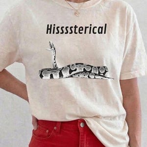 May include: A cream-colored short-sleeve t-shirt featuring a black and white graphic of a snake with its mouth open. The text "Hissssterical" is printed in black above the snake illustration. The shirt has a crew neckline and a relaxed fit.