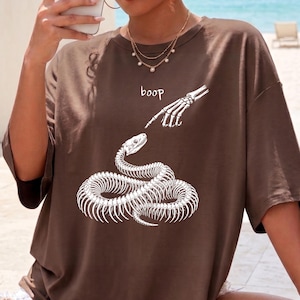 May include: A brown short-sleeved t-shirt featuring a white graphic print of a coiled snake skeleton being touched on the nose by a skeletal finger. The text "boop" is printed in white above the snake. The shirt has a crew neckline and loose fit.