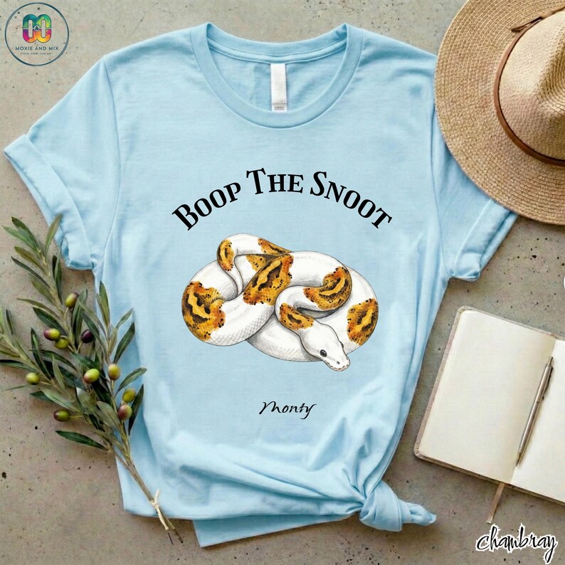 May include: Light blue t-shirt featuring a graphic of a white and orange snake with the text "Boop The Snoot" above it and the name "Monty" below. The shirt is knotted at the bottom.