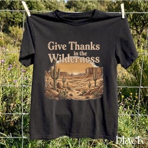May include: A black t-shirt featuring a graphic print of a desert landscape with cacti, rock formations, and a sunset. The text "Give Thanks in the Wilderness" is printed in a tan color above the illustration, which shows a sandy path.