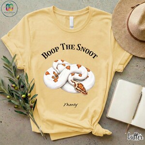 May include: Butter yellow t-shirt featuring a coiled white and orange snake illustration. The text "Boop The Snoot" is above the snake, with "Monty" below. A straw hat, notebook, and pen are visible.