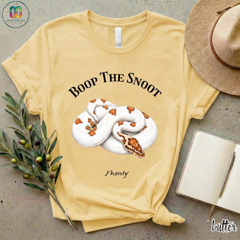 May include: Butter yellow t-shirt featuring a coiled white and orange snake illustration. The text "Boop The Snoot" is above the snake, with "Monty" below. A straw hat, notebook, and pen are visible.