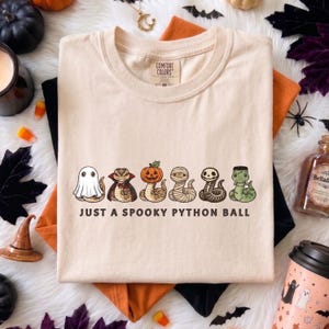 May include: A cream-colored short-sleeve t-shirt featuring a graphic of six cartoon snakes dressed as Halloween characters, including a ghost, vampire, pumpkin, mummy, skeleton, and Frankenstein. The text reads, JUST A SPOOKY PYTHON BALL.