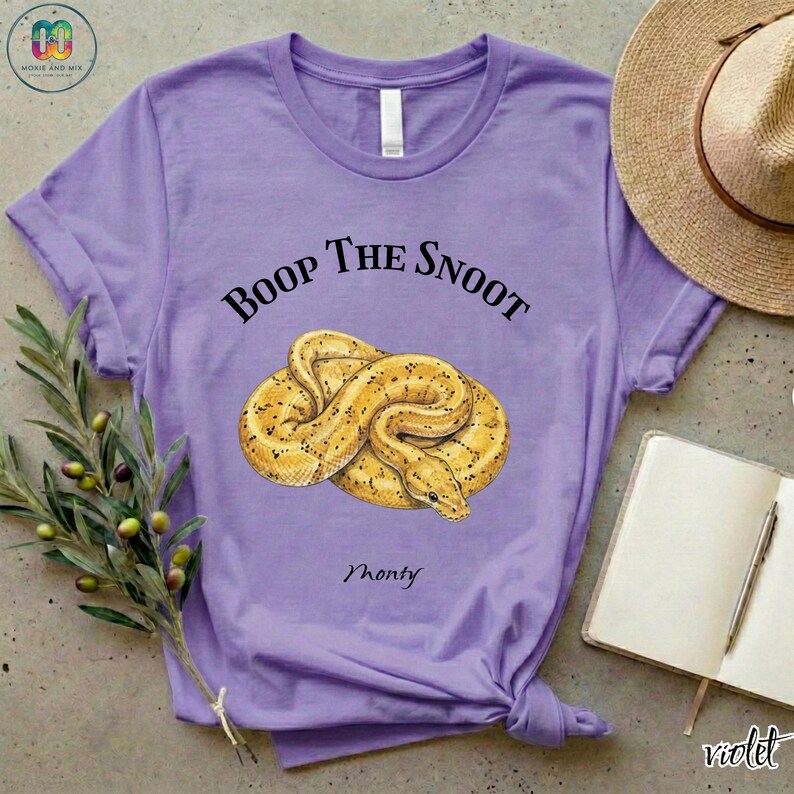 May include: A violet t-shirt with the text "Boop the Snoot" above a yellow snake illustration. The word "Monty" is below the snake. A straw hat, notebook, and olive branch are in the background.