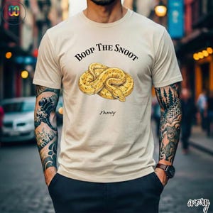May include: Ivory-colored t-shirt featuring a detailed illustration of a coiled yellow snake. The text "Boop The Snoot" is arched above the snake, with "Monty" written below. The shirt is a casual wear item.