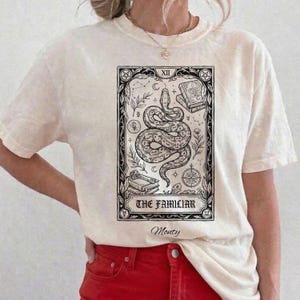 May include: A cream-colored short-sleeve t-shirt featuring a black ink-style graphic of a coiled snake, books, and celestial symbols inside a rectangular frame. The text reads THE FAMILIAR and Monty in a decorative script font below the frame.