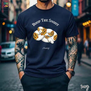 May include: Navy blue t-shirt featuring a white and yellow snake illustration with the text "Boop The Snoot" above and "Monty" below the snake. The shirt is a solid color.