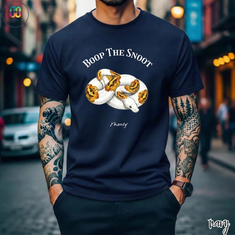 May include: Navy blue t-shirt featuring a white and yellow snake illustration with the text "Boop The Snoot" above and "Monty" below the snake. The shirt is a solid color.
