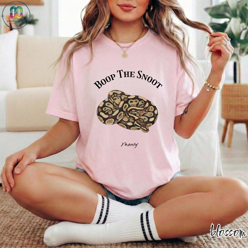 May include: A light pink t-shirt with the text "Boop The Snoot" above a graphic of a snake. The word "Monty" is below the snake graphic. The word "Blossom" is in the bottom right corner. The person is wearing white socks with black stripes.