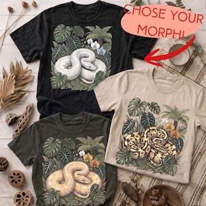 May include: Three short-sleeve t-shirts in black, beige, and dark gray, each featuring a graphic print of a coiled snake surrounded by tropical foliage and flowers. The text CHOSE YOUR MORPH! is printed in red above the black shirt.