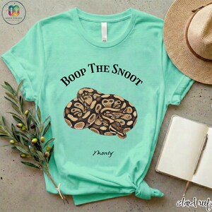 May include: A light teal t-shirt featuring a coiled snake illustration in shades of brown and tan. The text "Boop the Snoot" is above the snake, with "Monty" below. The shirt is knotted at the bottom.