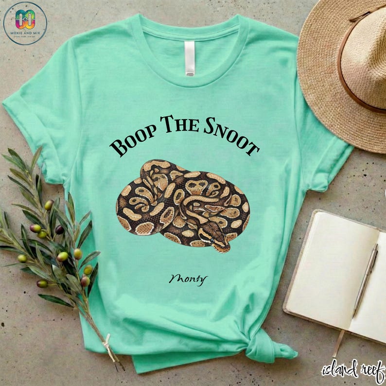 May include: A light teal t-shirt featuring a coiled snake illustration in shades of brown and tan. The text "Boop the Snoot" is above the snake, with "Monty" below. The shirt is knotted at the bottom.