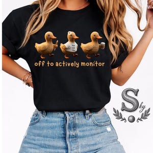 Puede incluir: Camiseta negra con tres patos de dibujos animados. Un pato sostiene un bolígrafo, otro lleva una camiseta que dice "You Got This", y el tercero sostiene una taza de café. El texto "off to actively monitor" está impreso debajo.
