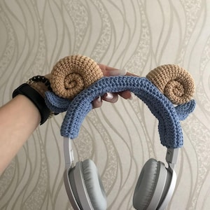 K&ouml;nnte beinhalten: Graue Kopfh&ouml;rer mit einem blauen, geh&auml;kelten Stirnband, verziert mit zwei beigefarbenen Schneckenhaus-Designs. Das handgefertigte Accessoire wird pr&auml;sentiert, um das einzigartige Design und die Handwerkskunst hervorzuheben.