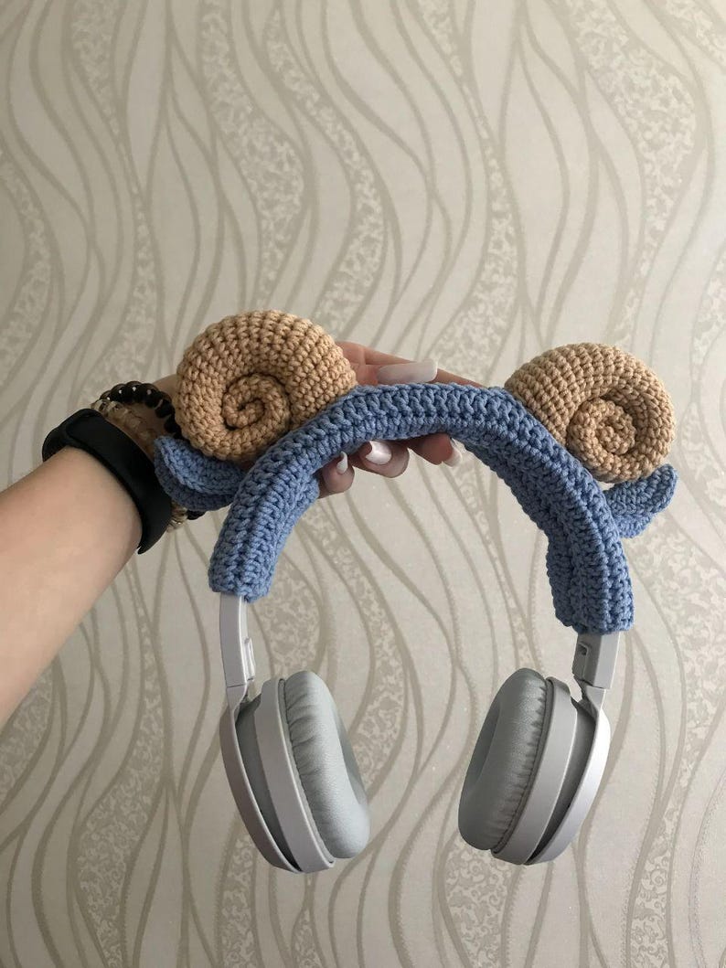 K&ouml;nnte beinhalten: Graue Kopfh&ouml;rer mit einem blauen, geh&auml;kelten Stirnband, verziert mit zwei beigefarbenen Schneckenhaus-Designs. Das handgefertigte Accessoire wird pr&auml;sentiert, um das einzigartige Design und die Handwerkskunst hervorzuheben.
