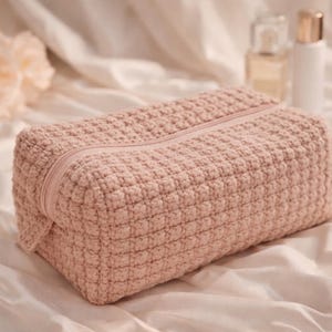 Puede incluir: Un neceser rectangular de ganchillo color rubor con cierre de cremallera a juego. La bolsa está hecha de un material texturizado y tejido y se encuentra sobre una superficie blanca y sedosa. Fondo suave con flores y botellas.
