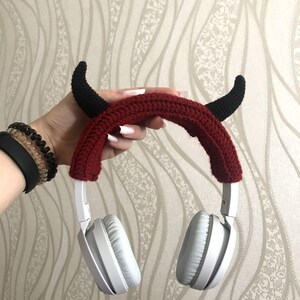 K&ouml;nnte beinhalten: Wei&szlig;e Kopfh&ouml;rer mit einem roten, geh&auml;kelten Stirnband mit schwarzen Teufelsh&ouml;rnern. Die Kopfh&ouml;rer haben ein gepolstertes Ohrmuscheldesign und werden von einer Hand gehalten. Das Stirnband ist dunkelrot.