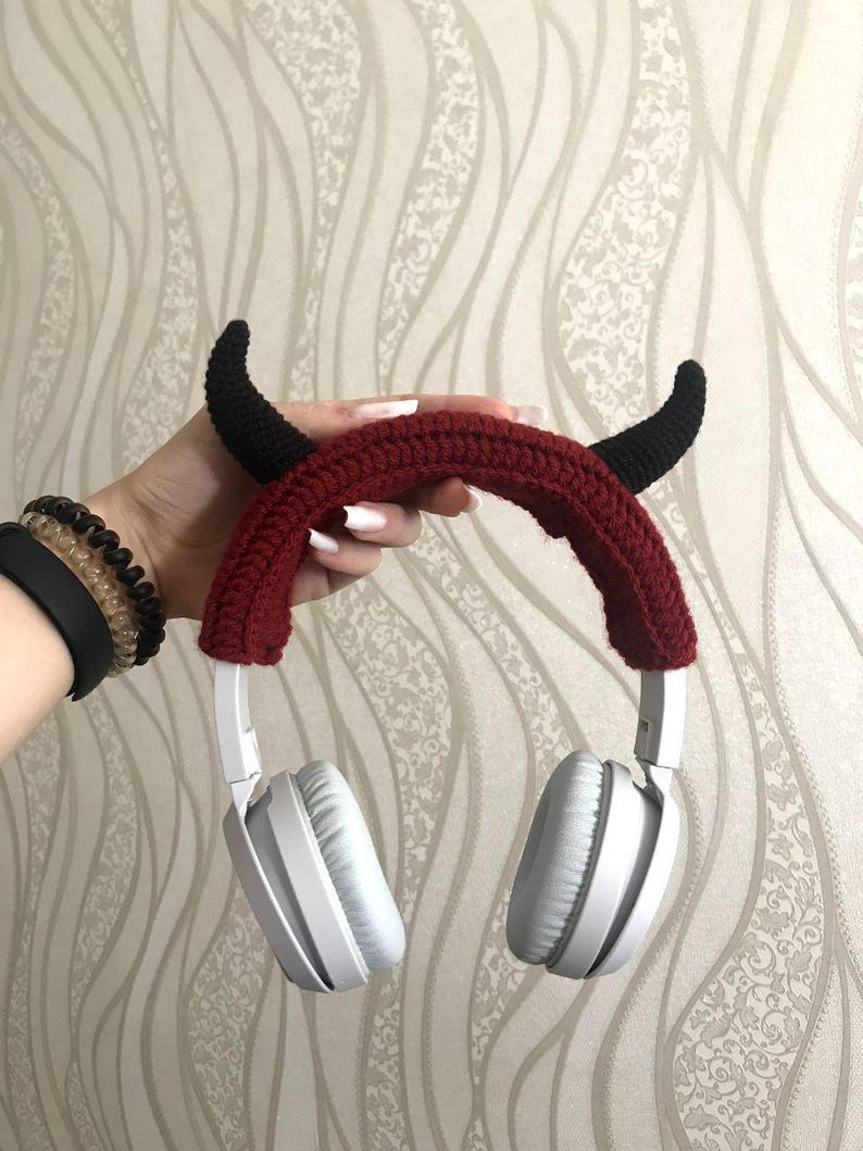 K&ouml;nnte beinhalten: Wei&szlig;e Kopfh&ouml;rer mit einem roten, geh&auml;kelten Stirnband mit schwarzen Teufelsh&ouml;rnern. Die Kopfh&ouml;rer haben ein gepolstertes Ohrmuscheldesign und werden von einer Hand gehalten. Das Stirnband ist dunkelrot.