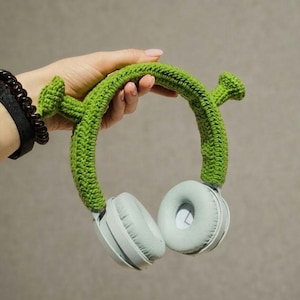 May include: Light green crochet cover for headphones featuring two protruding trumpet-shaped ears on the headband. The cover is made of textured yarn and fits over the top of light blue over-ear headphones with circular ear cups and padding.