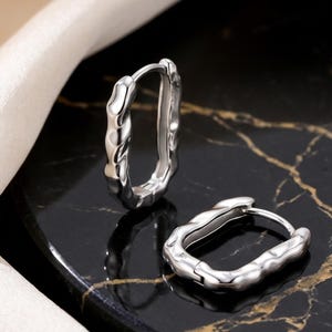 May include: A pair of silver oval hoop earrings with a textured, irregular design. The earrings are presented on a black and gold marble surface, with a white satin fabric in the background.